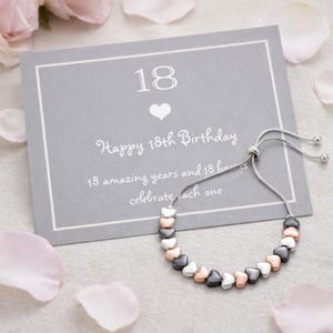 Peut inclure: Un bracelet argenté orné de perles en forme de cœur argentées, or rose et noires. Le bracelet est présenté avec une carte grise sur laquelle on peut lire "Joyeux 18e anniversaire" avec le chiffre 18 et un symbole de cœur. Des pétales de fleurs roses entourent la carte et le bracelet.