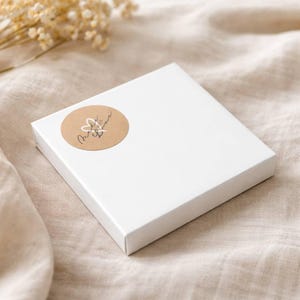 Peut inclure: Une bo&icirc;te cadeau carr&eacute;e en carton blanc avec un autocollant circulaire beige sur le coin sup&eacute;rieur gauche. L'autocollant pr&eacute;sente un contour de c&oelig;ur blanc dessin&eacute; &agrave; la main et le texte cursif noir Made with Love.