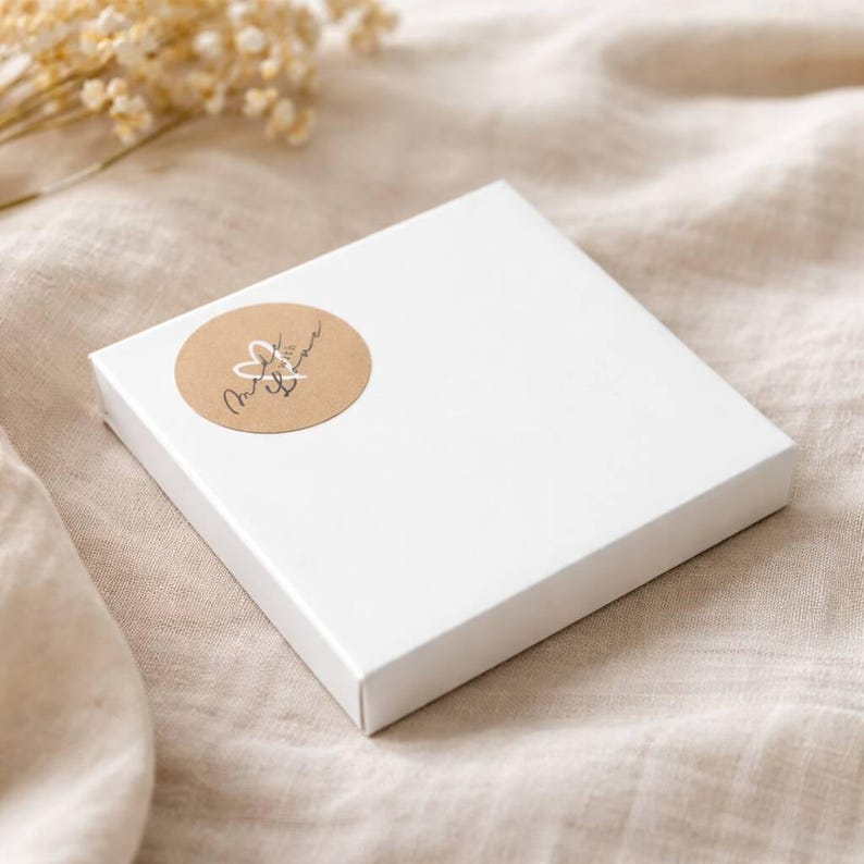 Peut inclure: Une bo&icirc;te cadeau carr&eacute;e en carton blanc avec un autocollant circulaire beige sur le coin sup&eacute;rieur gauche. L'autocollant pr&eacute;sente un contour de c&oelig;ur blanc dessin&eacute; &agrave; la main et le texte cursif noir Made with Love.