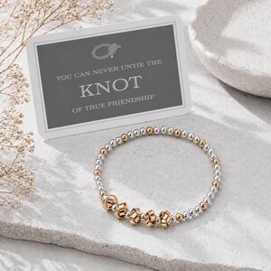 Può includere: Un braccialetto di perline con sfere metalliche alternate color argento e oro, con quattro perline centrali a forma di nodo dorato. Una carta rettangolare riporta il testo: YOU CAN NEVER UNTIE THE KNOT OF TRUE FRIENDSHIP.