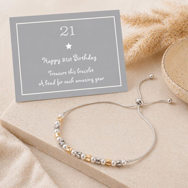 May include: A silver bracelet with a sliding clasp and a mix of gold, silver, and pearl beads. A gray card with the number "21" and the text "Happy 21st Birthday" is in the background. The card also reads "Treasure this bracelet, A bead for each amazing year."