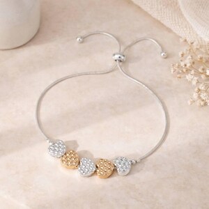 Peut inclure: Un bracelet ajustable de couleur argent&eacute;e orn&eacute; de cinq breloques en forme de c&oelig;ur dispos&eacute;es en alternance de couleurs argent et or. Les c&oelig;urs pr&eacute;sentent une surface textur&eacute;e et facett&eacute;e, avec un m&eacute;canisme de perle coulissante.