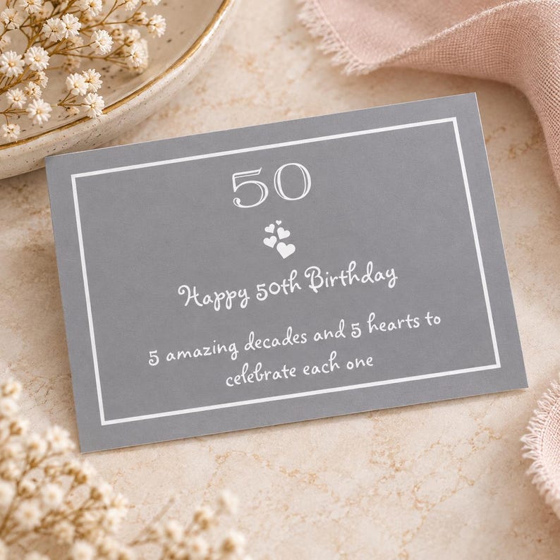 Peut inclure: Une carte rectangulaire grise avec une bordure blanche affichant le nombre 50, un groupe de cinq petits c&oelig;urs blancs et le texte : Happy 50th Birthday, 5 amazing decades and 5 hearts to celebrate each one, imprim&eacute; en police blanche manuscrite.