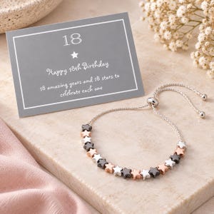 May include: A silver-tone adjustable slider bracelet featuring a series of small star-shaped beads in alternating metallic finishes of silver, rose gold, and dark grey, accompanied by a grey card printed with 18 and Happy 18th Birthday.