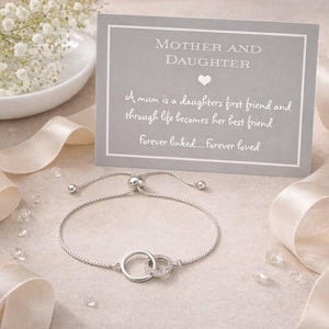 Puede incluir: Una pulsera ajustable de tono plateado con dos dijes circulares entrelazados, uno de los cuales está incrustado con pequeñas piedras transparentes. La pulsera se muestra con una tarjeta gris con el texto Mother and Daughter y un poema.