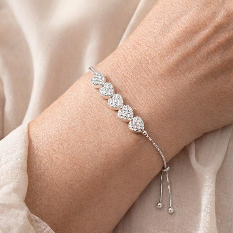 Peut inclure: Un bracelet argent&eacute; compos&eacute; de cinq breloques en forme de c&oelig;ur avec une surface textur&eacute;e et facett&eacute;e. Le bracelet est constitu&eacute; d'une fine cha&icirc;ne flexible et comprend un m&eacute;canisme coulissant r&eacute;glable avec deux petites perles argent&eacute;es aux extr&eacute;mit&eacute;s.