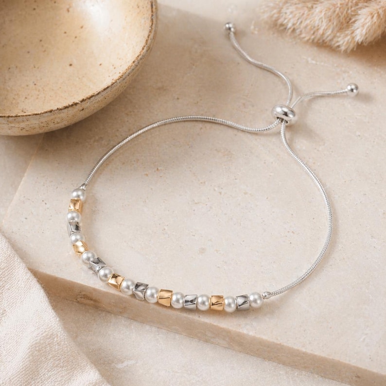 May include: Silver adjustable bracelet with a beaded design. The bracelet features a mix of silver, gold, and pearl-like beads. The adjustable closure allows for a customized fit. The bracelet is displayed on a neutral surface.
