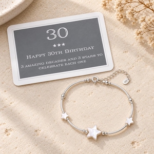 May include: A silver-toned bracelet featuring three white star-shaped charms and small round beads, accompanied by a rectangular card printed with the text 30, Happy 30th Birthday, and 3 amazing decades and 3 stars to celebrate each one.
