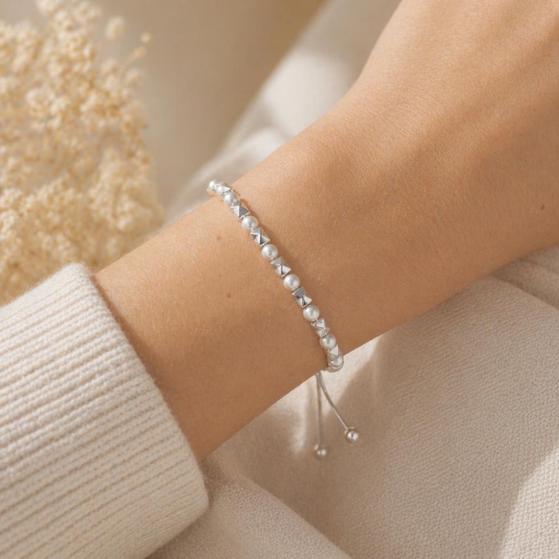May include: A delicate silver bracelet with small, round pearls and geometric silver accents. The bracelet is adjustable with a sliding closure and features two small silver beads at the ends. The jewelry is displayed on a person's wrist.