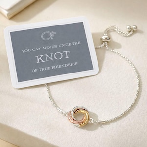 May include: A delicate adjustable bracelet featuring a central tri-color metal knot charm in silver, gold, and rose gold tones. It is displayed with a rectangular card printed with the text: YOU CAN NEVER UNTIE THE KNOT OF TRUE FRIENDSHIP.