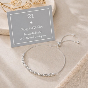 May include: A silver bracelet with a sliding clasp and a row of beads. A gray card with the number "21" and the text "Happy 21st Birthday, Treasure this bracelet, A bead for each amazing year" is in the background.