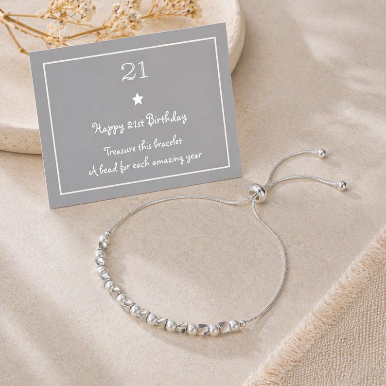 May include: A silver bracelet with a sliding clasp and a row of beads. A gray card with the number "21" and the text "Happy 21st Birthday, Treasure this bracelet, A bead for each amazing year" is in the background.