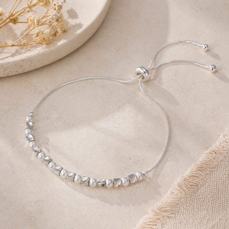 May include: A silver adjustable bracelet with a row of small, round, and square-shaped beads. The bracelet has a sliding clasp for easy adjustment. The beads are a light, pearlescent color, and the bracelet is set against a neutral background.