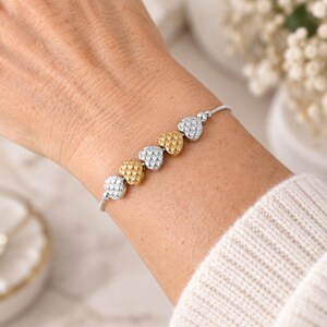 Peut inclure: Un bracelet d&eacute;licat compos&eacute; de cinq breloques en forme de c&oelig;ur alternant entre des tons argent&eacute;s et dor&eacute;s. Chaque c&oelig;ur pr&eacute;sente une surface textur&eacute;e et alv&eacute;ol&eacute;e. Le bracelet est constitu&eacute; d'une fine cha&icirc;ne m&eacute;tallique flexible.