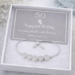 Peut inclure: Un bracelet r&eacute;glable argent&eacute; avec cinq breloques en forme de c&oelig;ur textur&eacute;, pr&eacute;sent&eacute; dans une bo&icirc;te cadeau blanche avec une carte grise portant l'inscription : 50, Happy 50th Birthday, 5 amazing decades and 5 hearts to celebrate each one.