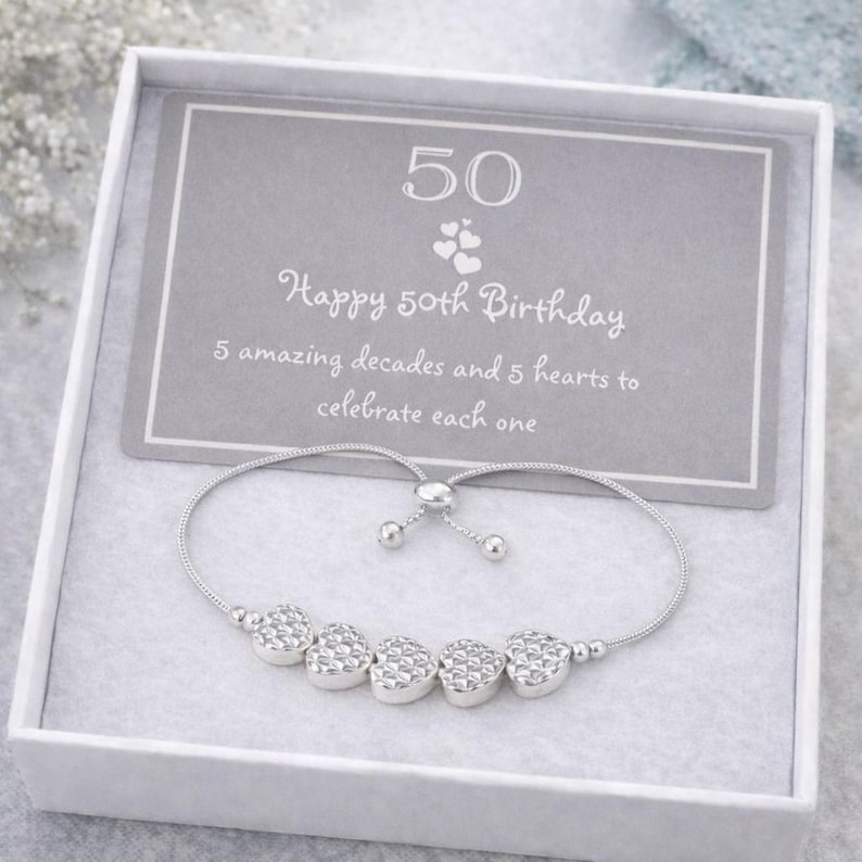 Peut inclure: Un bracelet r&eacute;glable argent&eacute; avec cinq breloques en forme de c&oelig;ur textur&eacute;, pr&eacute;sent&eacute; dans une bo&icirc;te cadeau blanche avec une carte grise portant l'inscription : 50, Happy 50th Birthday, 5 amazing decades and 5 hearts to celebrate each one.