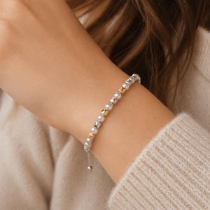 May include: A silver and gold beaded bracelet with small, round pearls and square gold accents. The bracelet is on a wrist, and the background is a neutral color.