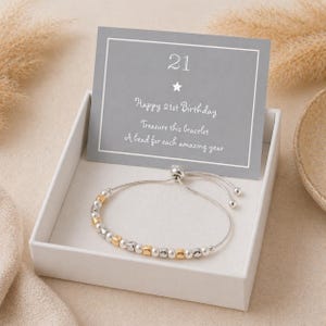 May include: A silver bracelet with gold and silver beads, presented in a white gift box. A gray card with the number "21" and the text "Happy 21st Birthday" is displayed behind the bracelet. The bracelet is a delicate piece of jewelry.