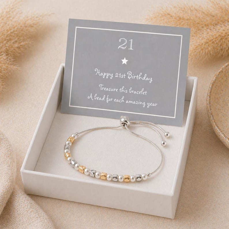 May include: A silver bracelet with gold and silver beads, presented in a white gift box. A gray card with the number "21" and the text "Happy 21st Birthday" is displayed behind the bracelet. The bracelet is a delicate piece of jewelry.