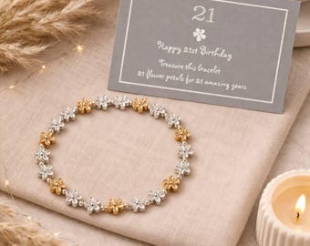 Könnte beinhalten: Ein Blumenarmband mit gold- und silberfarbenen blütenförmigen Gliedern wird auf einem beigen Tuch gezeigt. Eine graue Karte mit der Zahl 21 und dem Text "Happy 21st Birthday" befindet sich neben dem Armband. Eine brennende Kerze und dekoratives Weizen sind ebenfalls im Bild.
