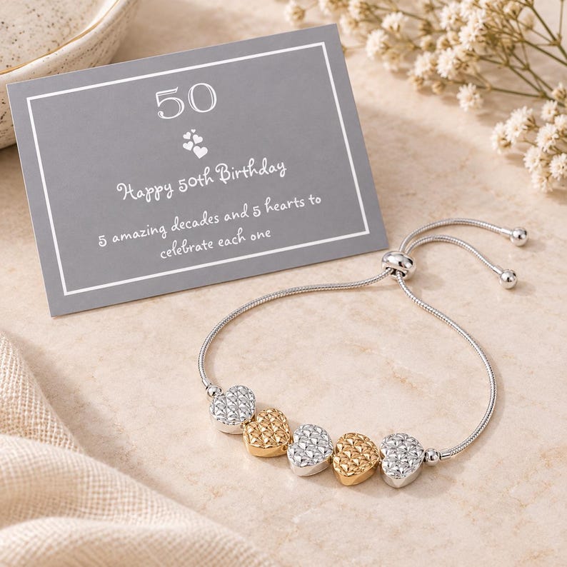 Peut inclure: Un bracelet coulissant r&eacute;glable argent&eacute; orn&eacute; de cinq breloques en forme de c&oelig;ur textur&eacute;, alternant entre les tons argent et or. Une carte rectangulaire grise accompagne le bracelet, portant l'inscription 50 Happy 50th Birthday 5 amazing decades and 5 hearts to celebrate each one.