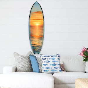 May include: A wooden surfboard with a sunset scene painted on it. The surfboard is hanging on a white wall with a couch and pillows in front of it.