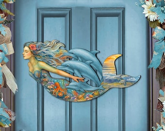 May include: A wooden mermaid and dolphin wall decoration. The mermaid has long flowing hair and a flower in her hair. The dolphins are light blue. The mermaid's tail features an ocean scene with a sunset.