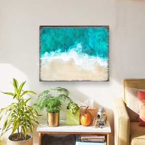 Puede incluir: Arte mural cuadrado que representa una vista a&eacute;rea de una playa. La obra de arte presenta agua turquesa del oc&eacute;ano que se encuentra con una orilla arenosa. La pieza est&aacute; enmarcada en madera oscura y r&uacute;stica. El arte se exhibe sobre una mesa auxiliar con plantas y decoraci&oacute;n.