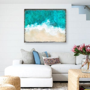 Puede incluir: Una obra de arte enmarcada que representa una vista a&eacute;rea de una playa. La obra presenta agua turquesa, olas blancas y una playa arenosa. El marco es de color marr&oacute;n oscuro. La obra se exhibe sobre un sof&aacute; blanco con cojines decorativos.