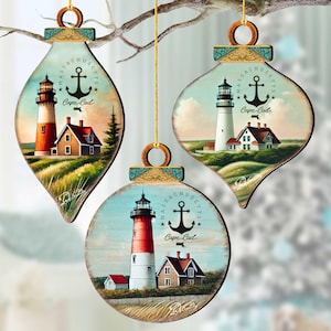 May include: Three wooden Christmas ornaments with lighthouse scenes. Each ornament features a different lighthouse design with the words "Massachusetts" and "Cape Cod". The ornaments are hanging from a tree branch.