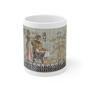 May include: White ceramic mug with a colorful image of an Egyptian pharaoh sitting on a throne. The pharaoh is wearing a golden crown and a white robe. Two figures are standing behind the pharaoh, one is holding a fan and the other is holding a lotus flower. The background is a beige papyrus-like texture with hieroglyphs and a depiction of lotus flowers.