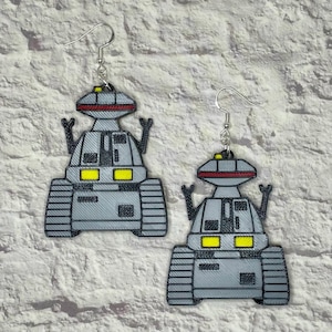 May include: A pair of silver robot earrings with yellow and red accents. The robots have a rounded body with a rectangular head and two arms raised in the air. The earrings are hanging from silver hooks.