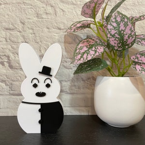 May include: A white and black wooden bunny figurine with a black top hat and a black mustache. The bunny is standing on a black surface in front of a white brick wall.
