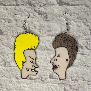 May include: A pair of earrings featuring cartoon characters from the animated television series Beavis and Butt-Head. The earrings are made of plastic and are painted in bright colors. The left earring features Beavis with his signature yellow hair and the right earring features Butt-Head with his brown hair.