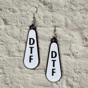 May include: A pair of white dangle earrings with black lettering that reads "DTF" on each earring.