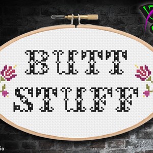 May include: A white cross-stitch embroidery hoop with the words "But First, Stuff" in black lettering. The letters have decorative flourishes. Two small pink and green flowers are on either side of the text.