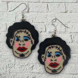 May include: A pair of dangle earrings featuring a stylized face with black hair, a pale beige complexion, blue eyeshadow, pink cheeks, and bright red lips. The face has black stitched details across the forehead and chin, suspended from silver-toned hooks.