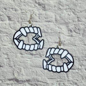 May include: A pair of white acrylic earrings shaped like a set of vampire fangs. The earrings are on a white brick background.