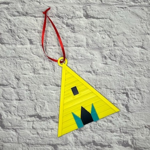 May include: A yellow triangular ornament with a black rectangle and black and teal shapes. The ornament has a red ribbon for hanging. The background is a white brick wall.