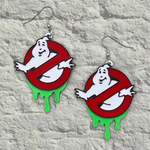 May include: A pair of Ghostbusters-themed earrings. Each earring features a white ghost figure inside a red no-ghost logo, with green slime dripping from the bottom. The earrings have silver-colored fish hook ear wires.