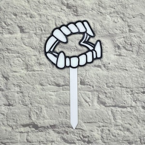 May include: A white and black vampire teeth-shaped cake topper. The teeth are outlined in black and have a white background. The topper is attached to a white stick, perfect for Halloween or vampire-themed parties.