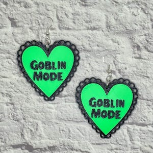 May include: A pair of green heart-shaped earrings with black lace trim and the text "Goblin Mode" printed in black.