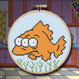 May include: A cross-stitch embroidery hoop with a cartoon image of a goldfish with three eyes. The goldfish is orange and swimming in blue water with green seaweed. The hoop is on a kitchen counter with cabinets in the background.