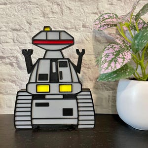 May include: A gray and black robot toy with yellow lights and a red stripe on the head. The robot has a track-like base and is standing on a black surface.