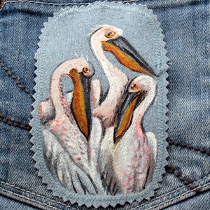 May include: A hand-painted patch featuring three pelicans. The birds have white feathers, pink accents, and orange and black beaks. The patch is on a light blue background with a scalloped edge, set against a denim pocket.