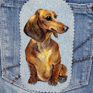 May include: A brown and tan dachshund dog is painted on a blue denim patch. The patch is sewn onto a pair of blue denim jeans.