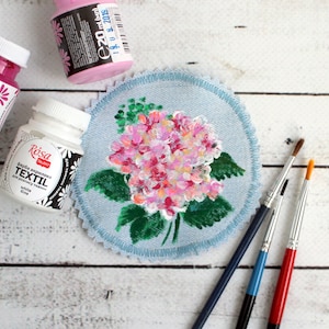 Op de afbeelding: Een close-up van een blauwe stoffen cirkel met een geschilderde roze en witte hortensia. Op de voorgrond liggen drie kwasten. De kwasten zijn rood, blauw en zwart. Op de achtergrond staan drie verfpotjes. De verfpotjes zijn roze, wit en rood. De verfpotjes zijn gelabeld met de tekst "Rósa TALENT фарба акрилова TEXTIL для розпису тканин white 20 ml (мл) 19.0 4...2019 -"