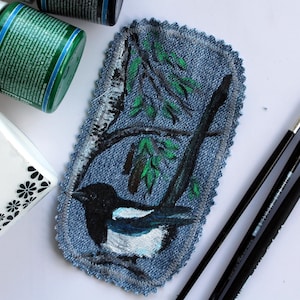 May include: A rectangular denim patch with scalloped edges featuring a hand-painted magpie perched on a branch. The bird has black and white plumage, and the background depicts a tree with green leaves and a white birch trunk painted on the blue fabric.