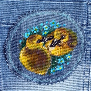 May include: A close-up of a denim patch with a hand-painted design of two yellow ducklings nestled amongst blue forget-me-not flowers.