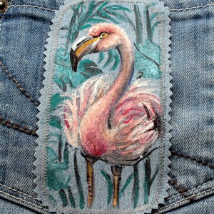 Pode incluir: Um patch retangular com um desenho de flamingo pintado. O flamingo é rosa com um bico amarelo e está em um fundo verde e turquesa. O patch é costurado em um fundo de ganga.